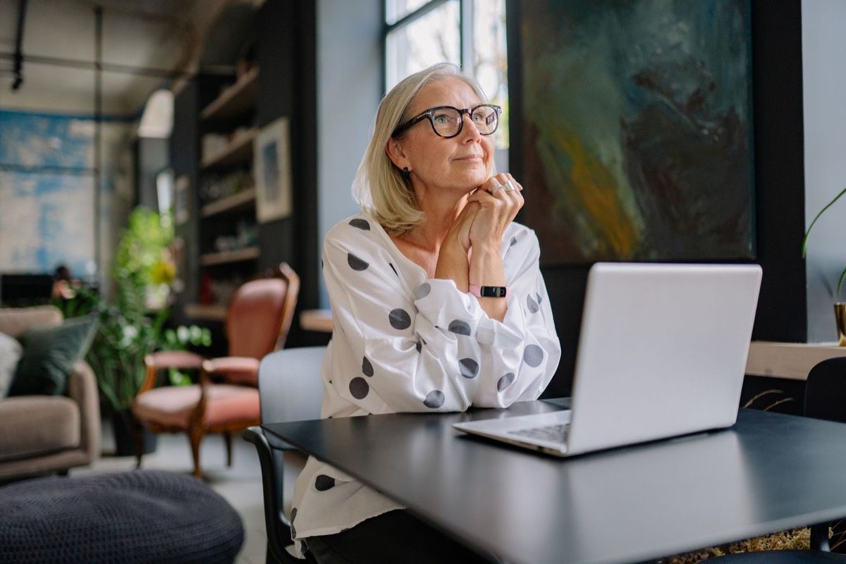 Woman in her late 50s with silver hair and glasses sitting at a desk with her laptop, hands clasped under her chin in a thoughtful evaluating pose
