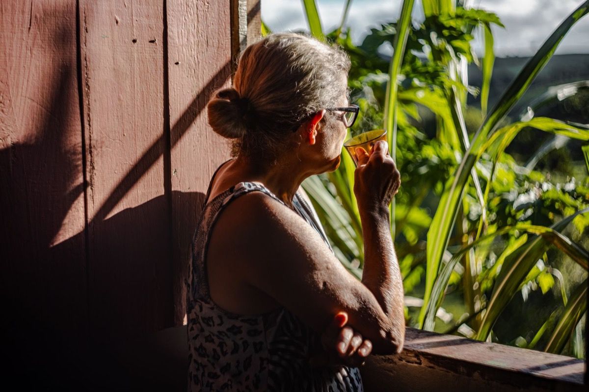 Woman over 50 sipping coffee alone on a sunny balcony, looking out toward greenery
