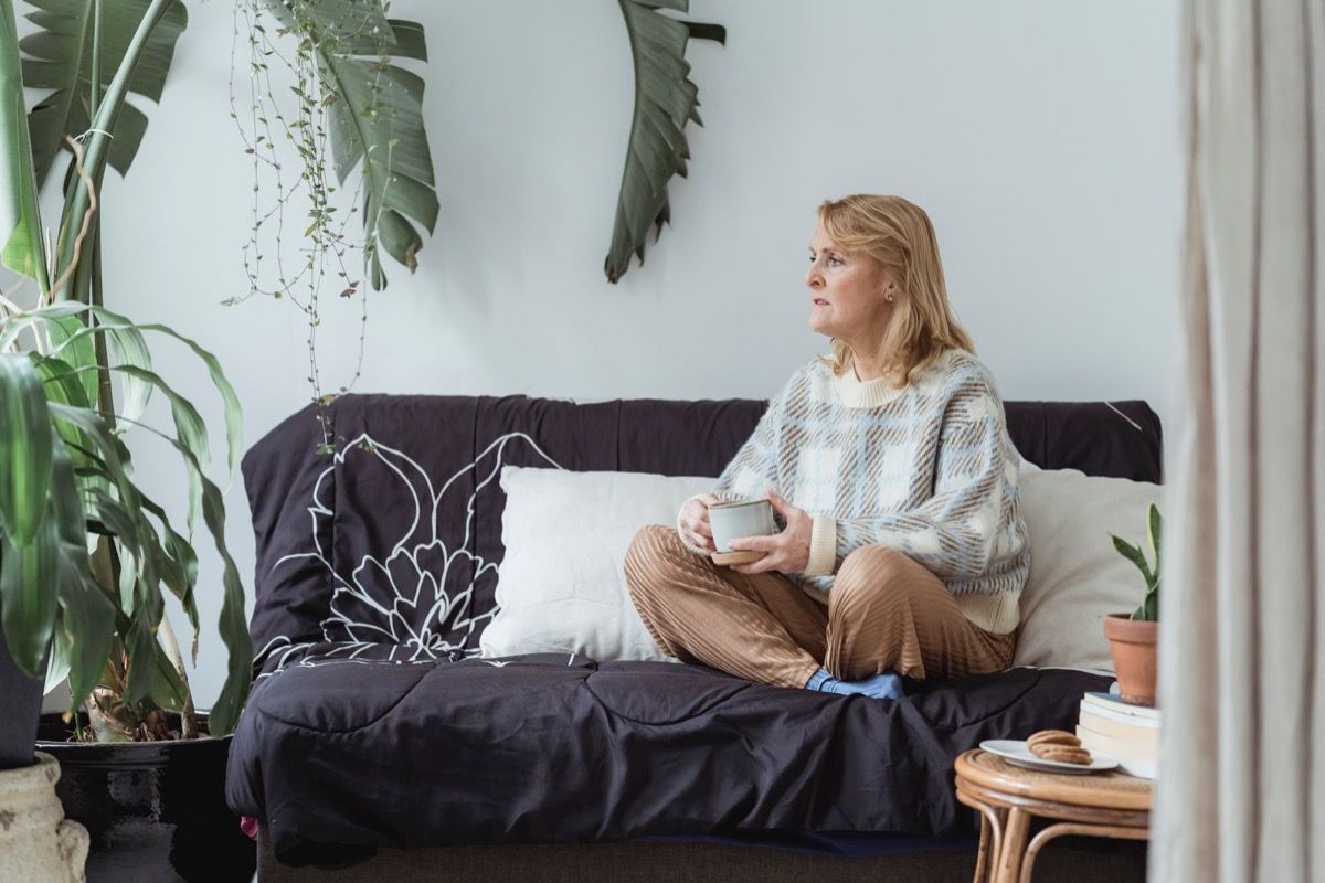 Woman in her 50s sitting on a sofa holding a warm drink, looking thoughtfully toward a window