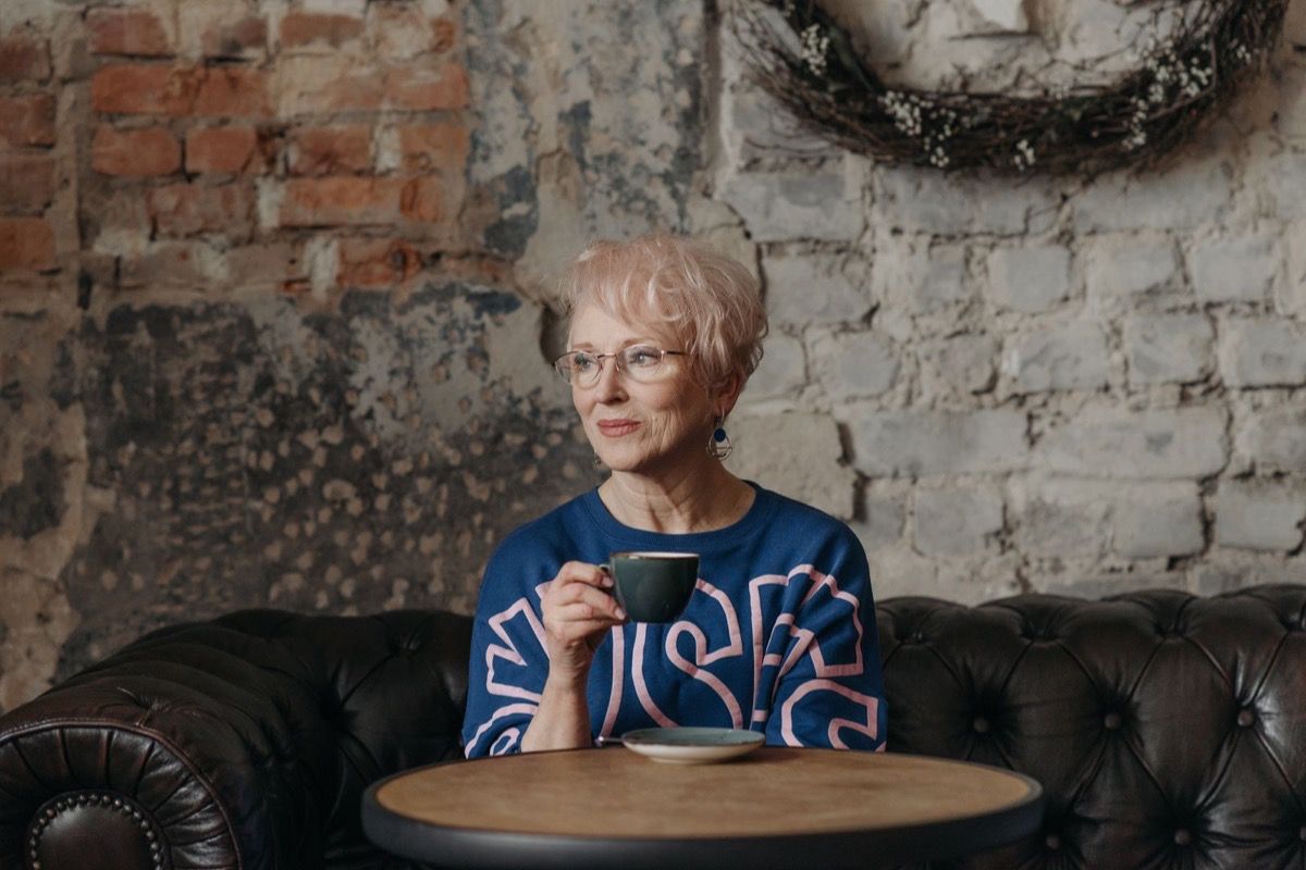 Woman in her 60s with short hair and glasses holding a coffee cup in a cozy cafe, looking away with quiet confidence