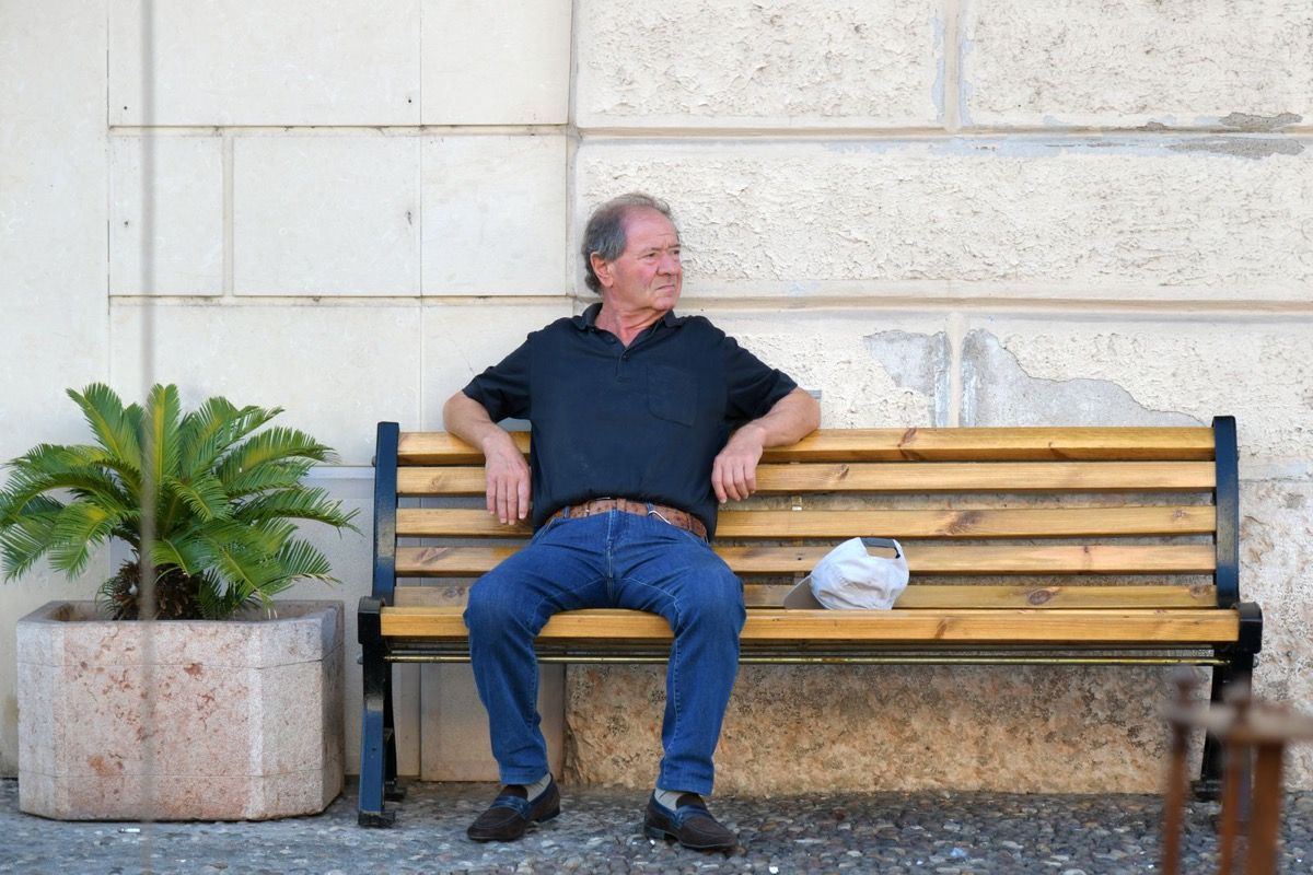 Man in his 70s sitting on a town bench in the sun, looking sideways with quiet interest