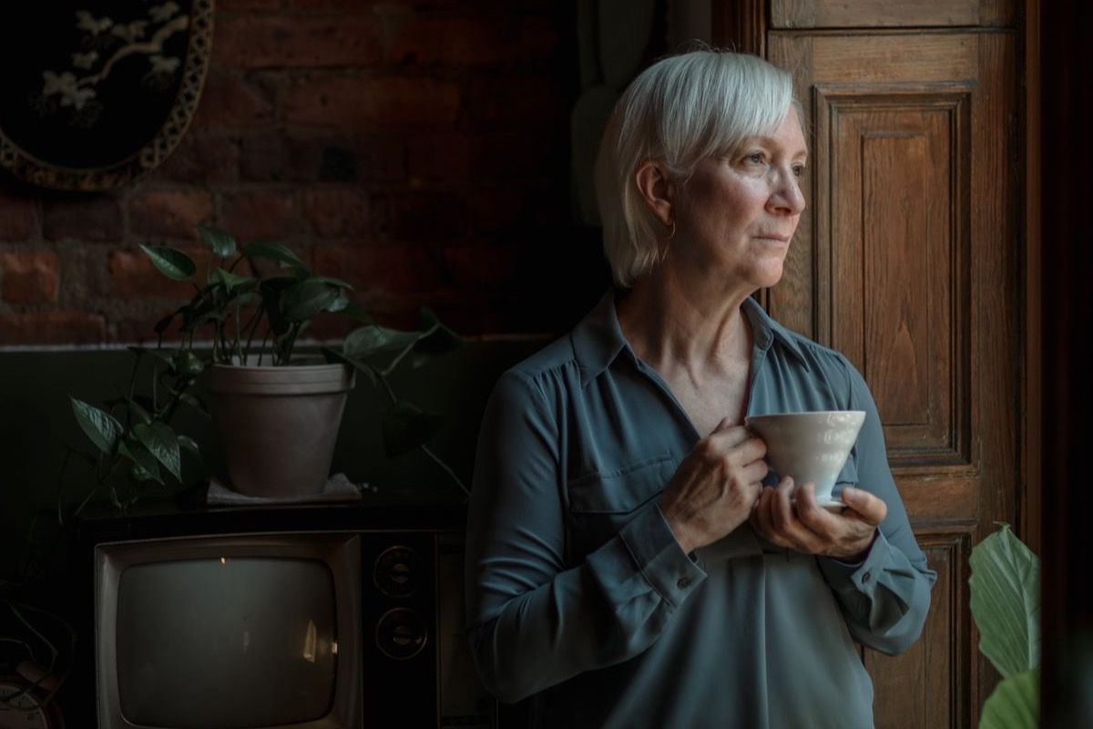 Woman over 50 holding a cup of coffee and gazing thoughtfully toward a window in a warm home setting