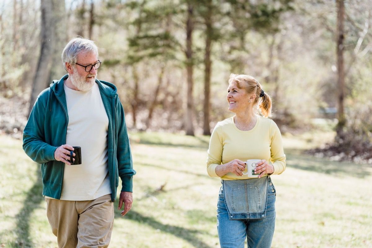 Two adults over 50 walking together through a sunny park holding coffee mugs, relaxed and engaged in conversation