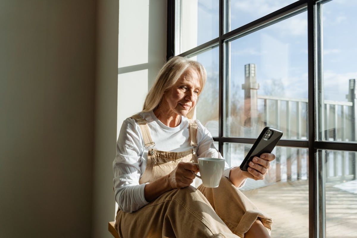 Woman over 50 holding a phone and coffee while reading a message by a bright window