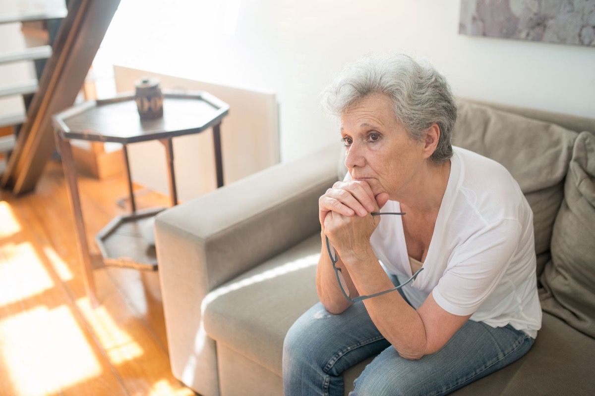 Woman over 60 sitting on a sofa holding her glasses, pausing in thought