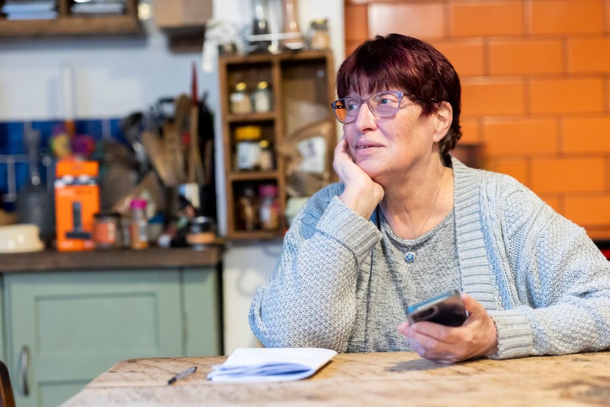 Woman in her 60s holding a smartphone at her kitchen table, gazing away thoughtfully