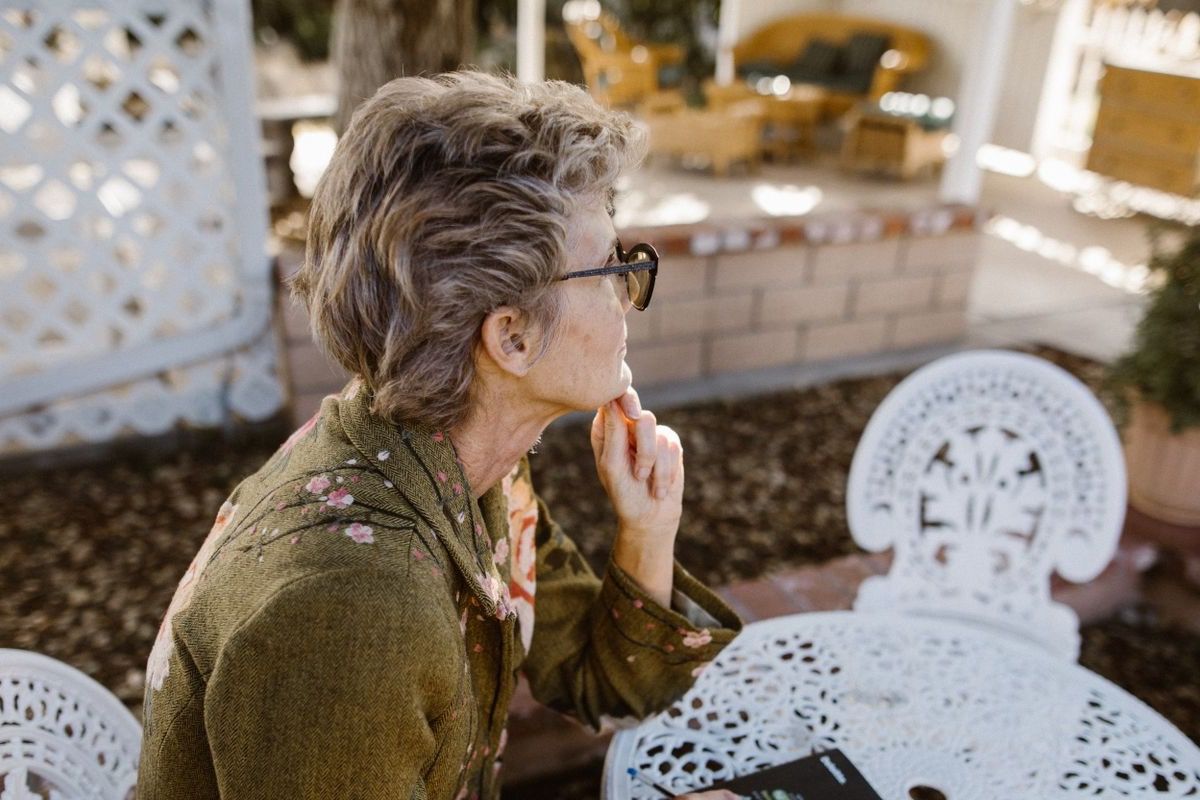 Woman over 60 with grey hair sitting at a garden table in warm light, hand near chin, looking thoughtfully into the distance