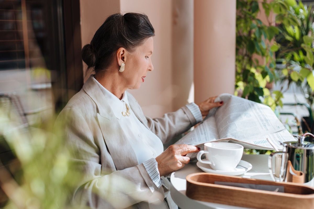 Mature woman reading a newspaper alone at an outdoor cafe with coffee, looking content and at ease