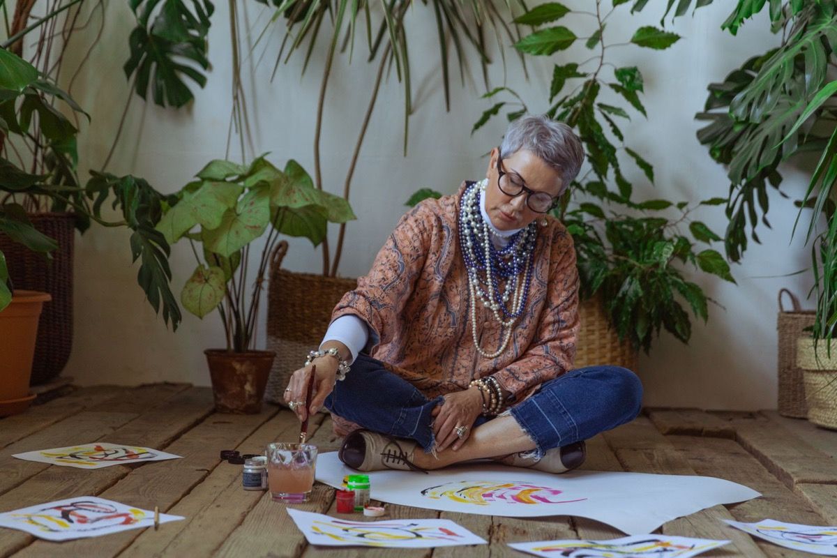 A woman over 50 sitting on the floor painting watercolors alone, absorbed in her own creative activity surrounded by houseplants
