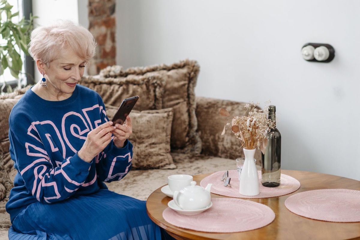Woman over 60 reviewing smartphone settings on a sofa with a teapot and cup on the table
