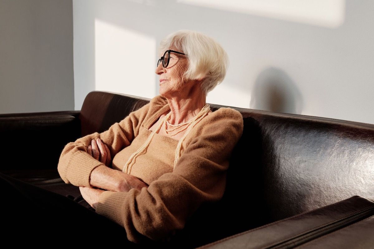 Older woman with white hair sitting on a leather sofa with arms crossed, looking away thoughtfully in natural light