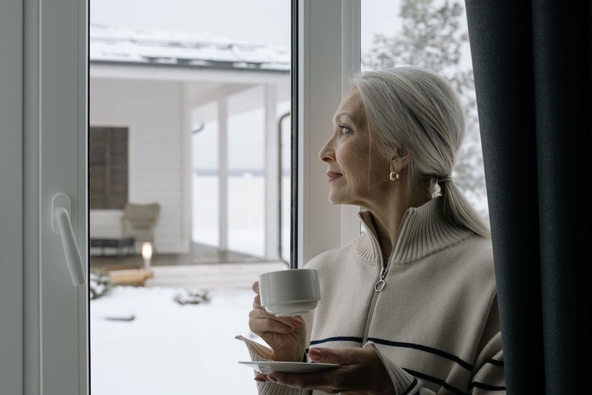 Woman over 60 with silver hair holding a coffee cup by a large window, gazing outward with a calm contemplative expression