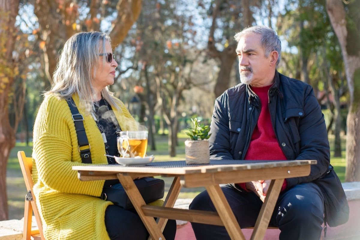 A man and woman in their 50s talking across a wooden table at an outdoor park cafe on a sunny day