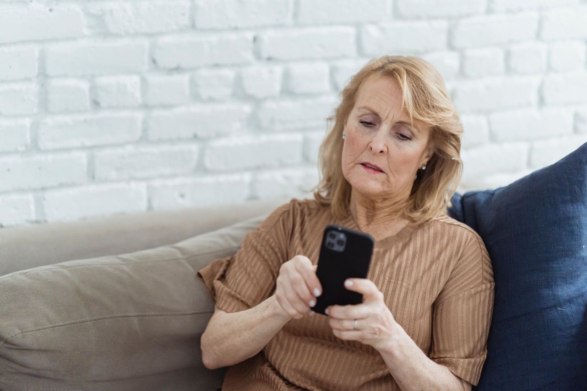 Woman in her late 50s looking pensively at her smartphone while sitting on a sofa at home
