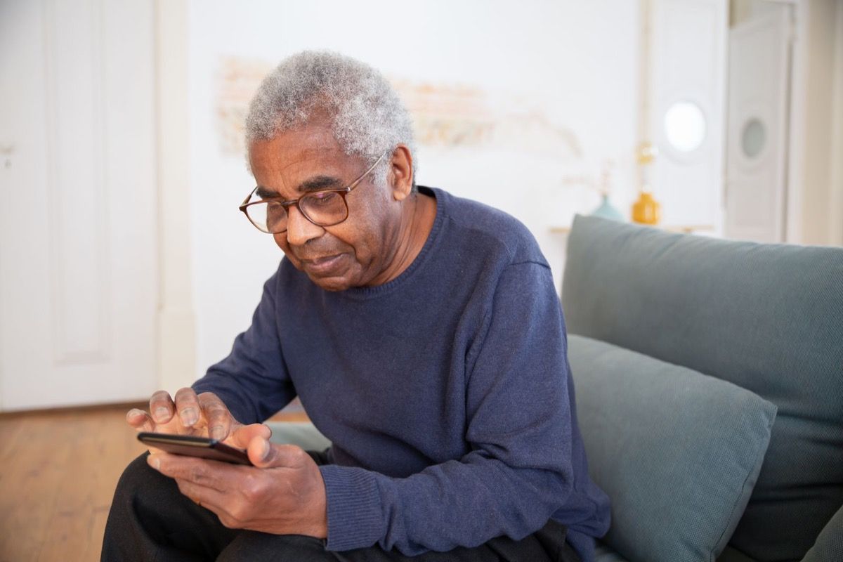 Man over 70 in a blue sweater sitting on a sofa, looking at his smartphone with a focused, contemplative expression